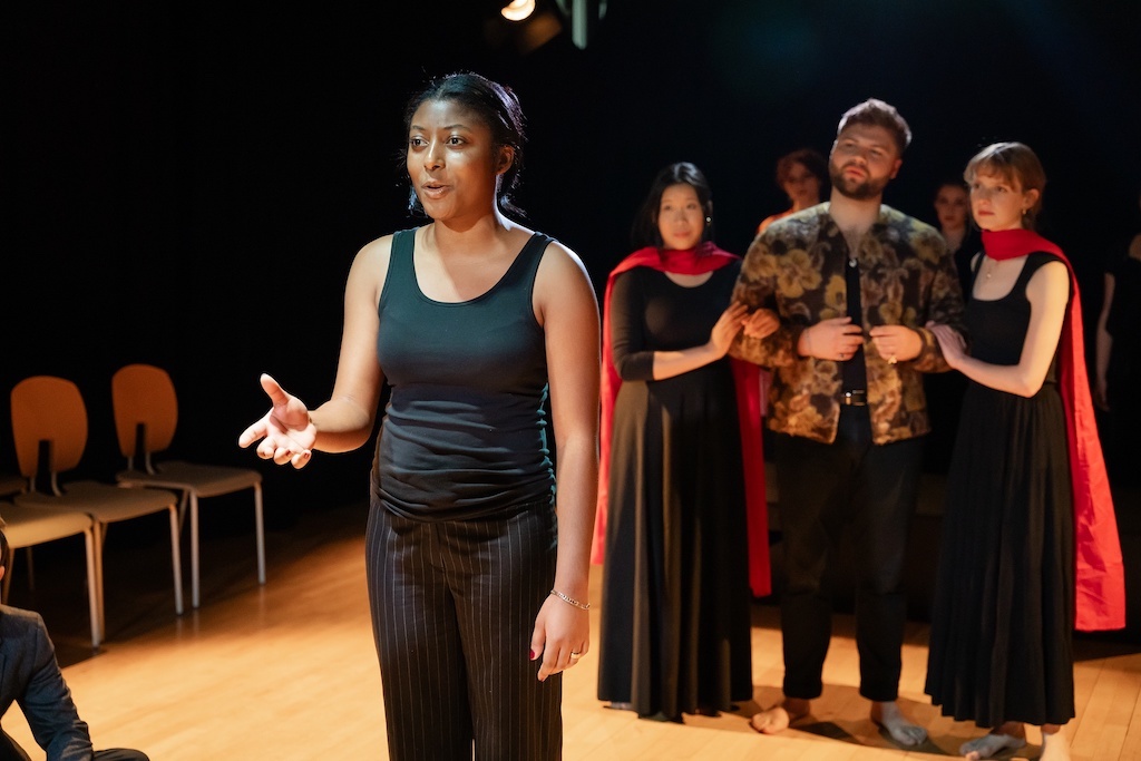 Actor stands with right hand stretched out in mid-speech while three other actors stand behind her, focused on her performance.